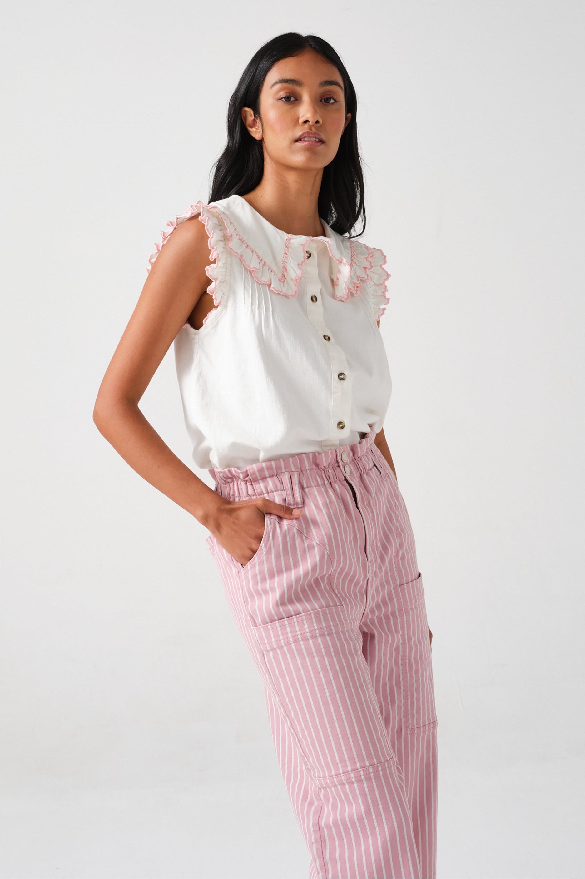 A woman wearing a short sleeve blouse with a frill collar and embroidered details in ecru and dusty rose colors, paired with striped pants.