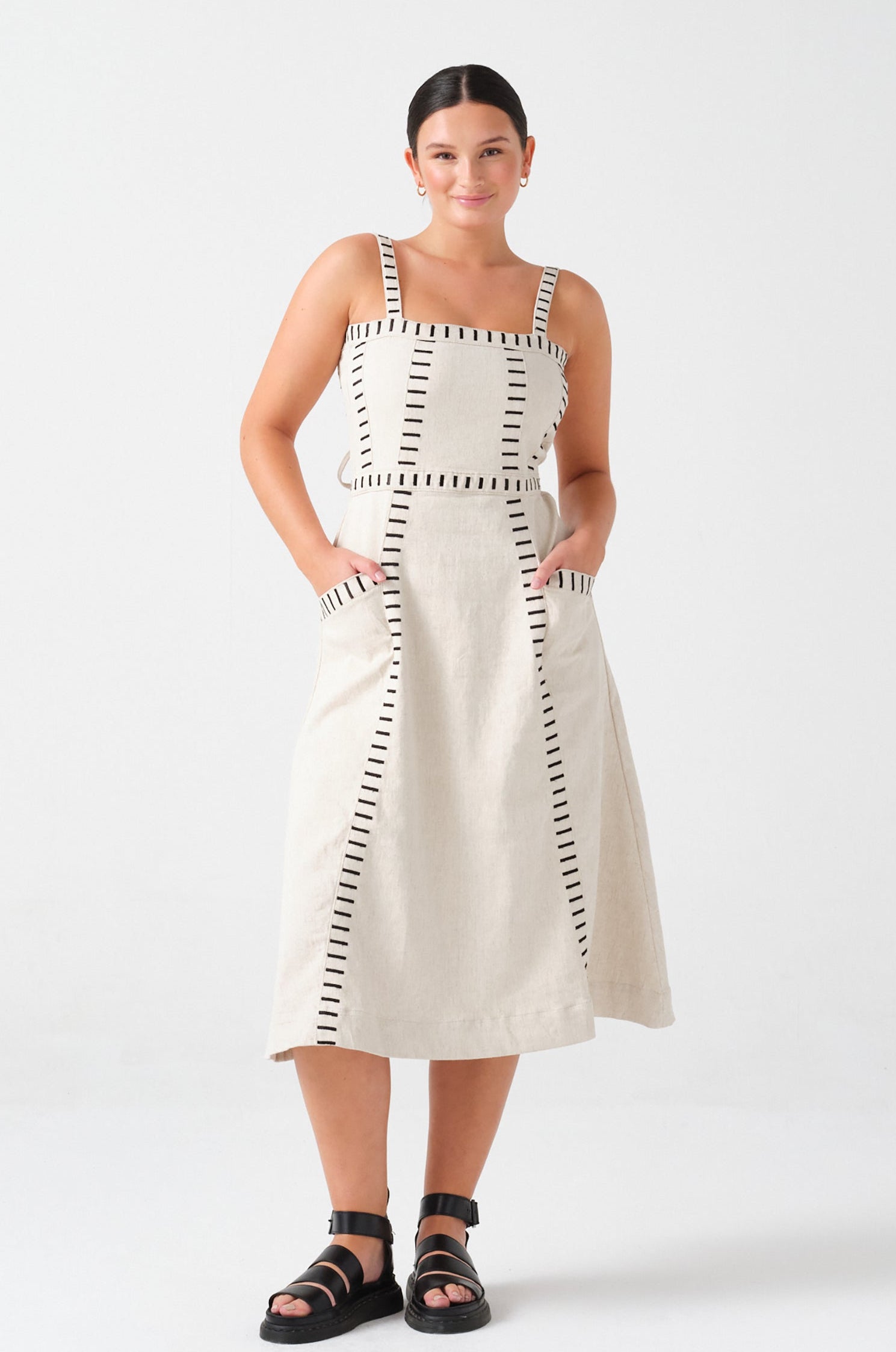 A woman wearing an ecru midi denim dress with black stitch details, a square neckline, front pockets, and an open, lace-up back on a white background