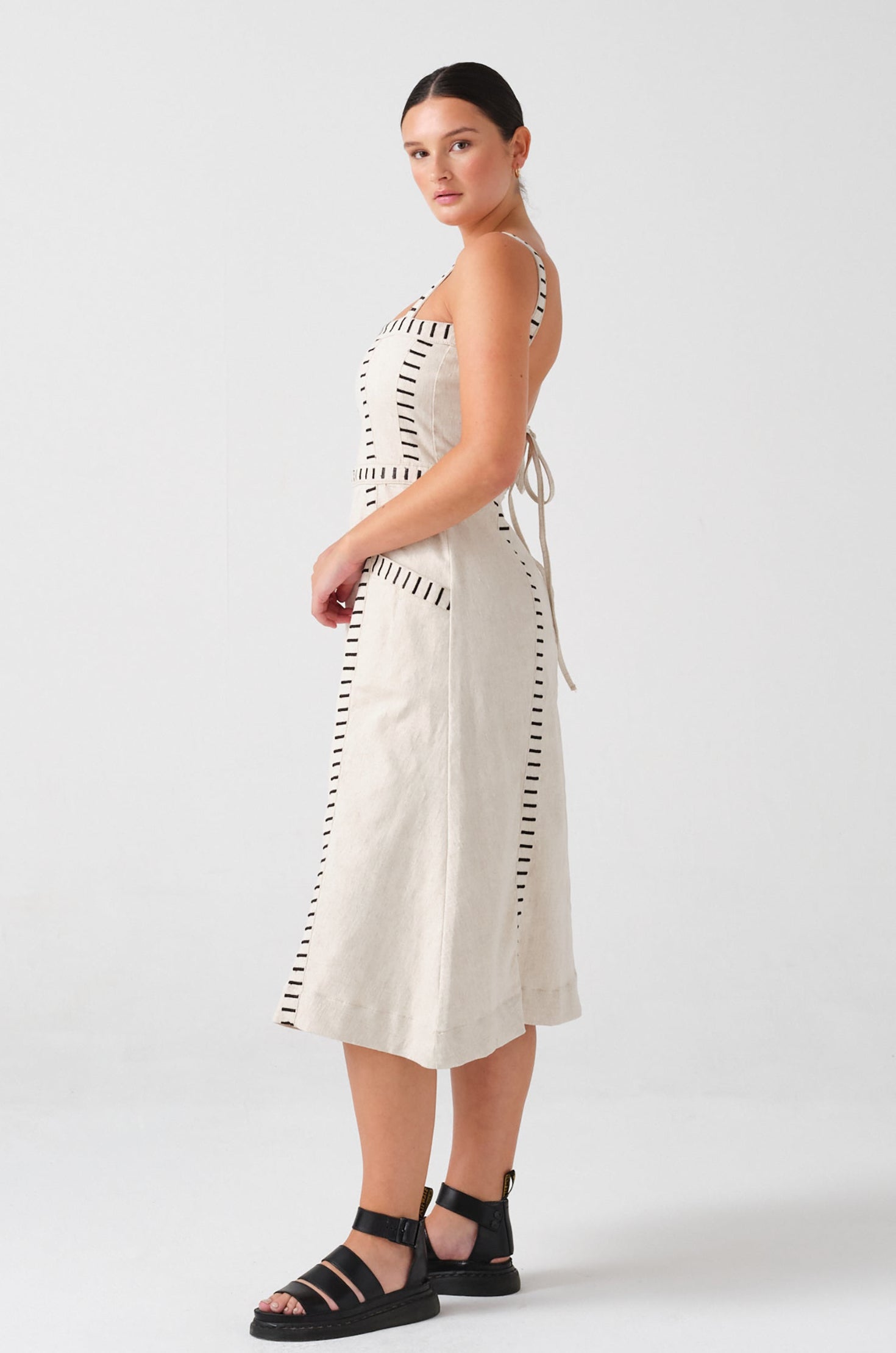 A woman wearing an ecru midi denim dress with black stitch details, a square neckline, front pockets, and an open, lace-up back on a white background