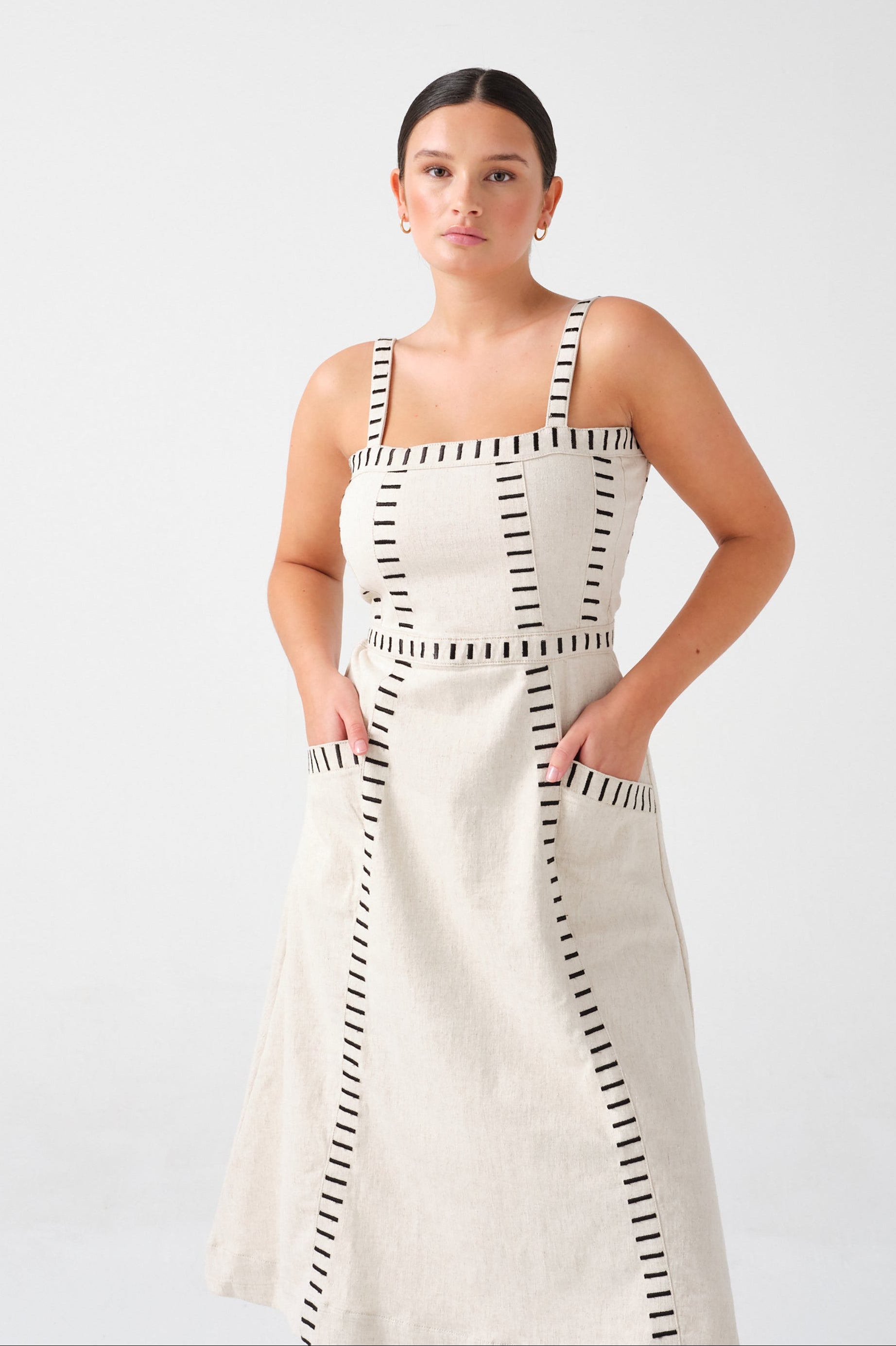 A woman wearing an ecru midi denim dress with black stitch details, a square neckline, front pockets, and an open, lace-up back on a white background