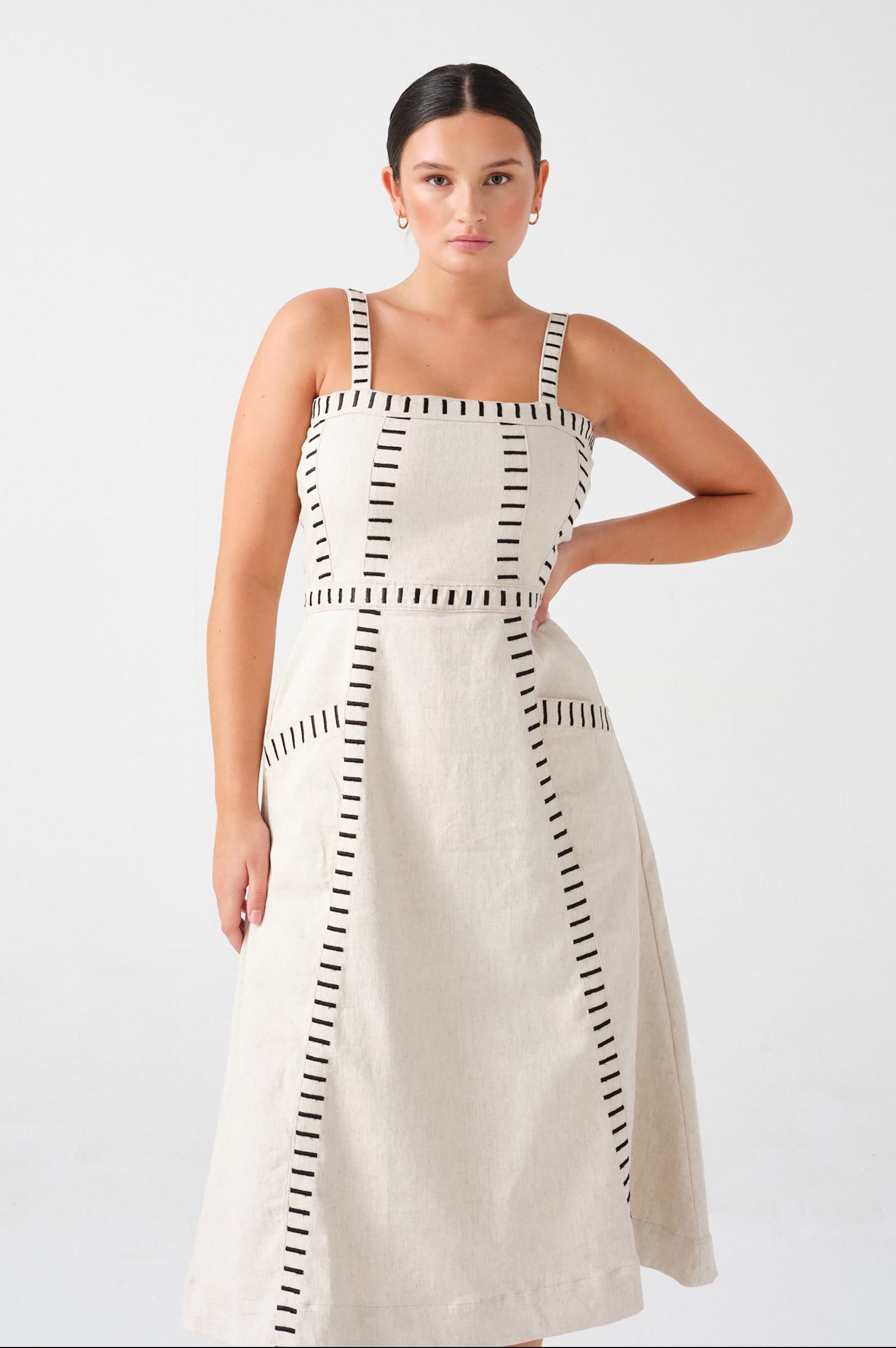 A woman wearing an ecru midi denim dress with black stitch details, a square neckline, front pockets, and an open, lace-up back on a white background