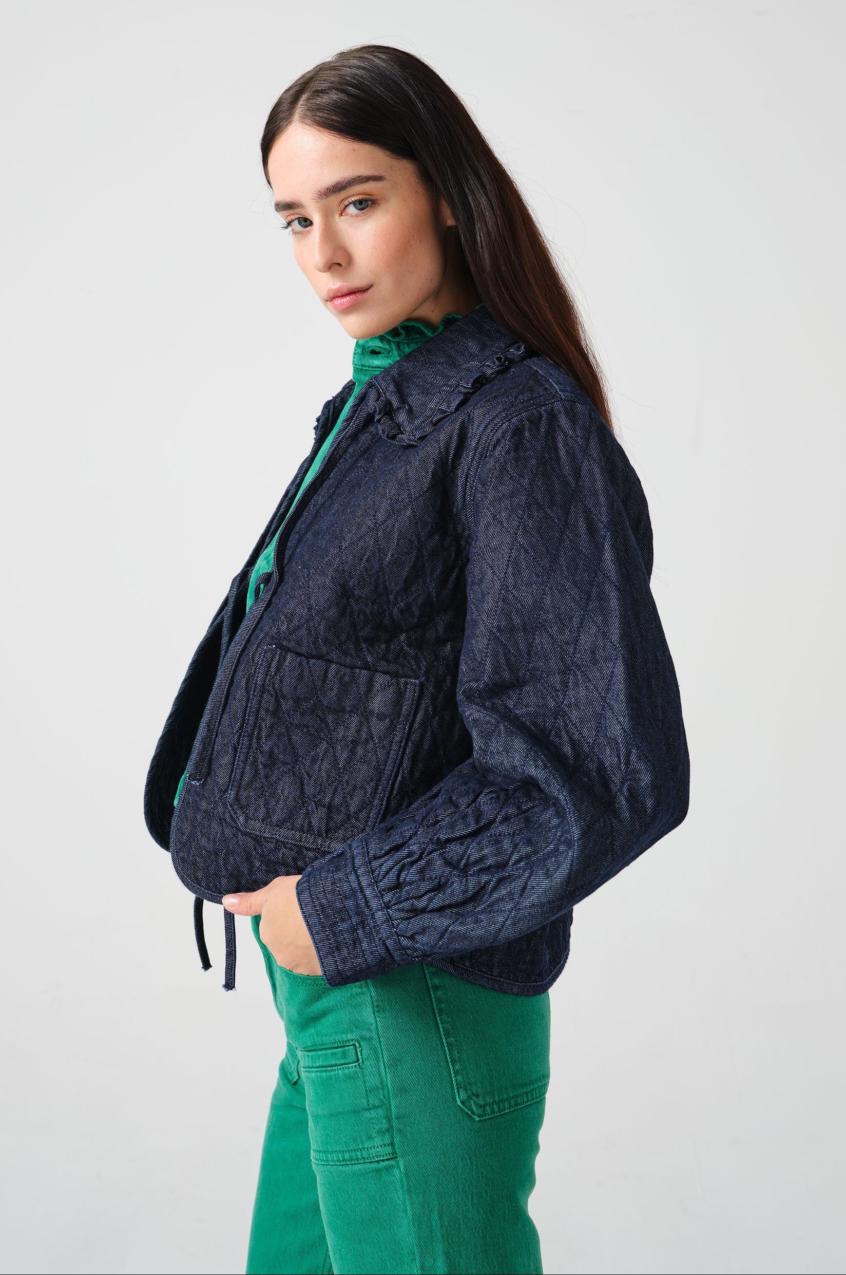 Woman wearing a dark blue denim quilted jacket with a slightly frilled collar and tie closures with a green denim shirt and green denim jeans
