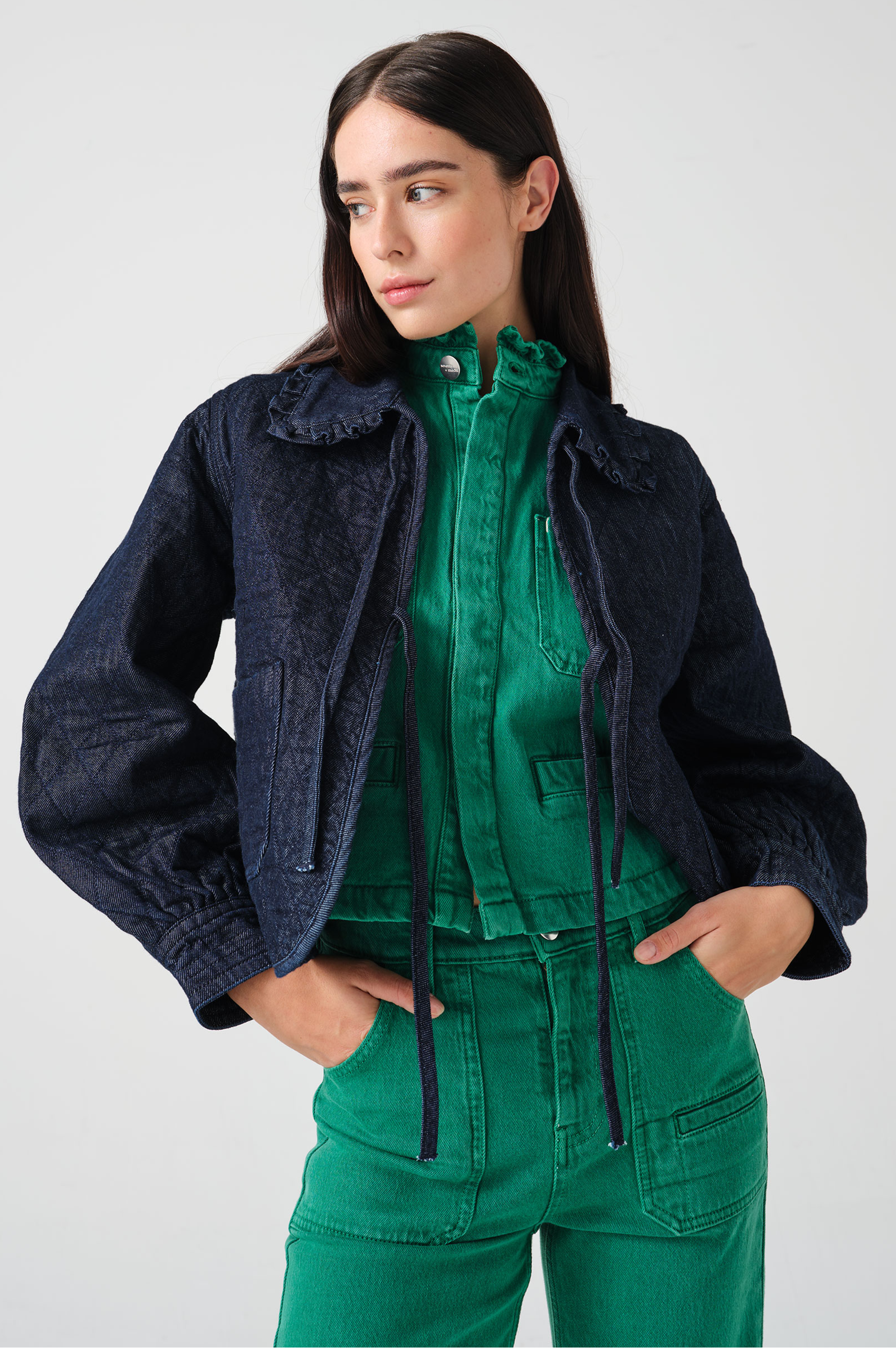 Woman wearing a dark blue denim quilted jacket with a slightly frilled collar and tie closures with a green denim shirt and green denim jeans