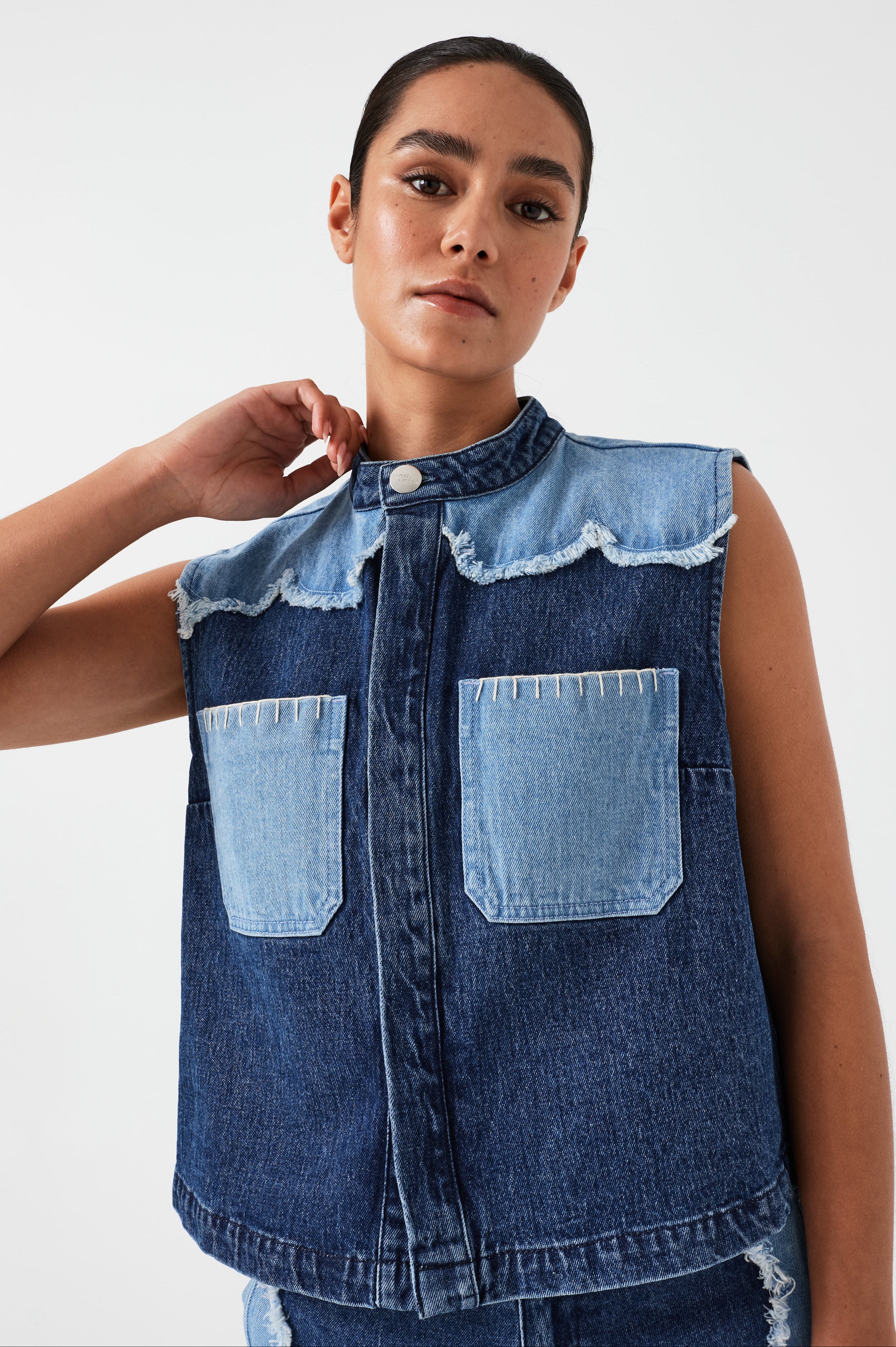 woman wearing a dark and light blue denim vest/ waistcoat with scallop frayed details and our signature white blanket stitching on two large patch pockets with blue jeans with scallop frayed details against a white backdrop
