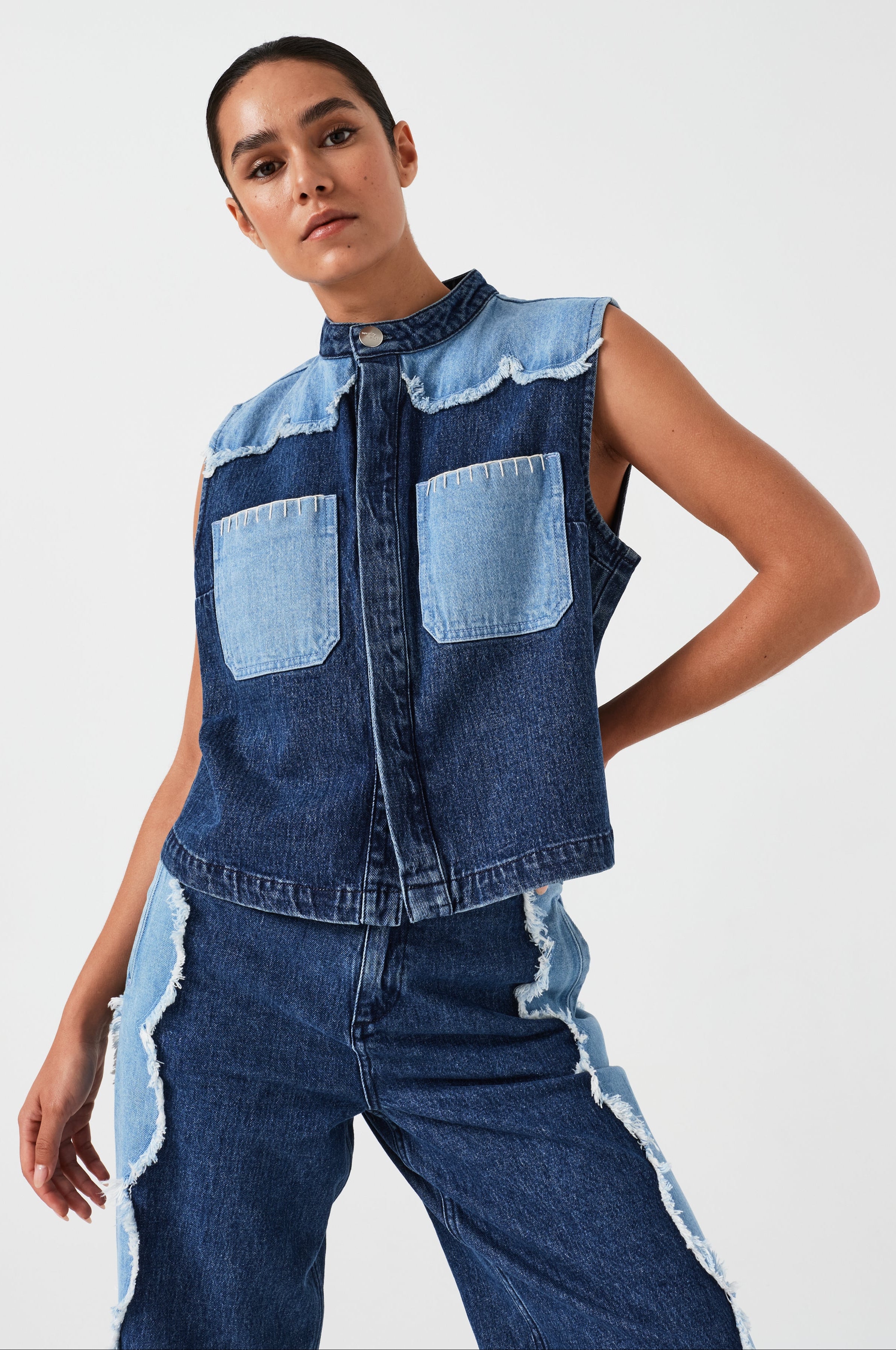 woman wearing a dark and light blue denim vest/ waistcoat with scallop frayed details and our signature white blanket stitching on two large patch pockets with blue jeans with scallop frayed details against a white backdrop