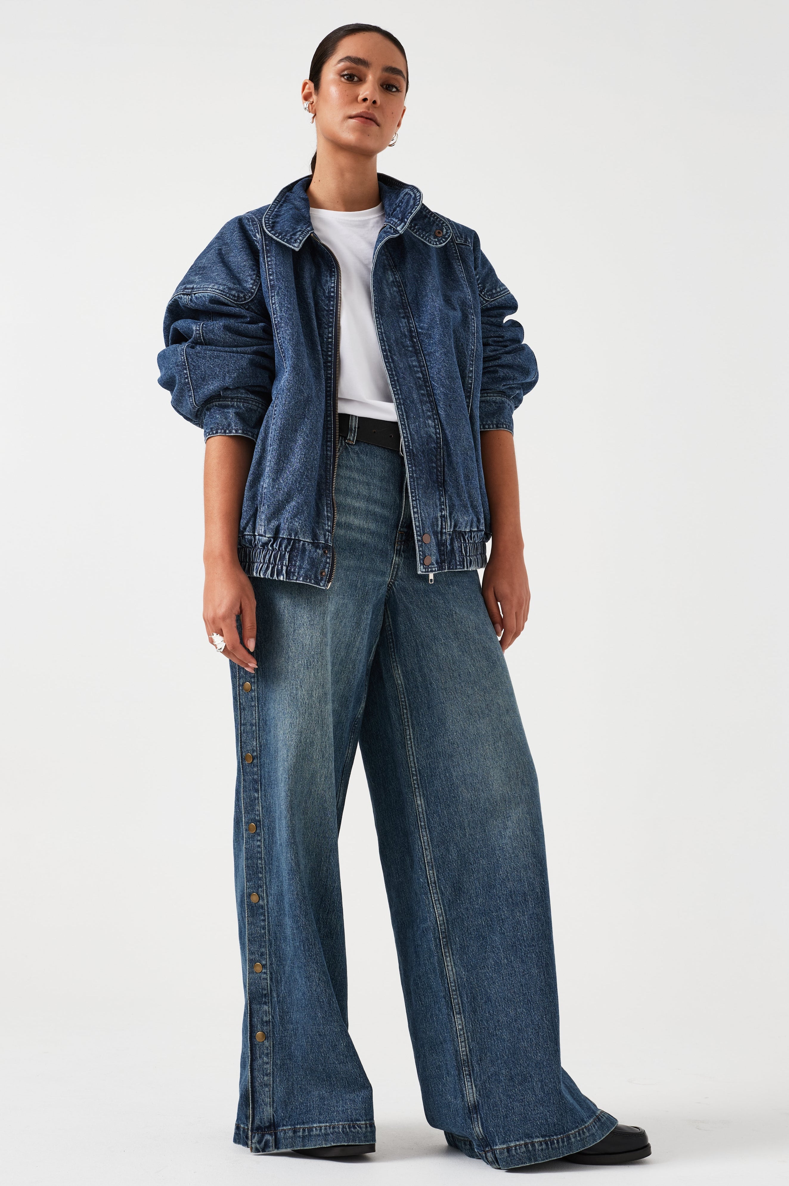 A woman wearing an oversized padded and seamed blue denim retro style bomber jacket with a round neckline, zip, and button closures at the top, bottom, and collar with a white top and blue jeans against a white backdrop