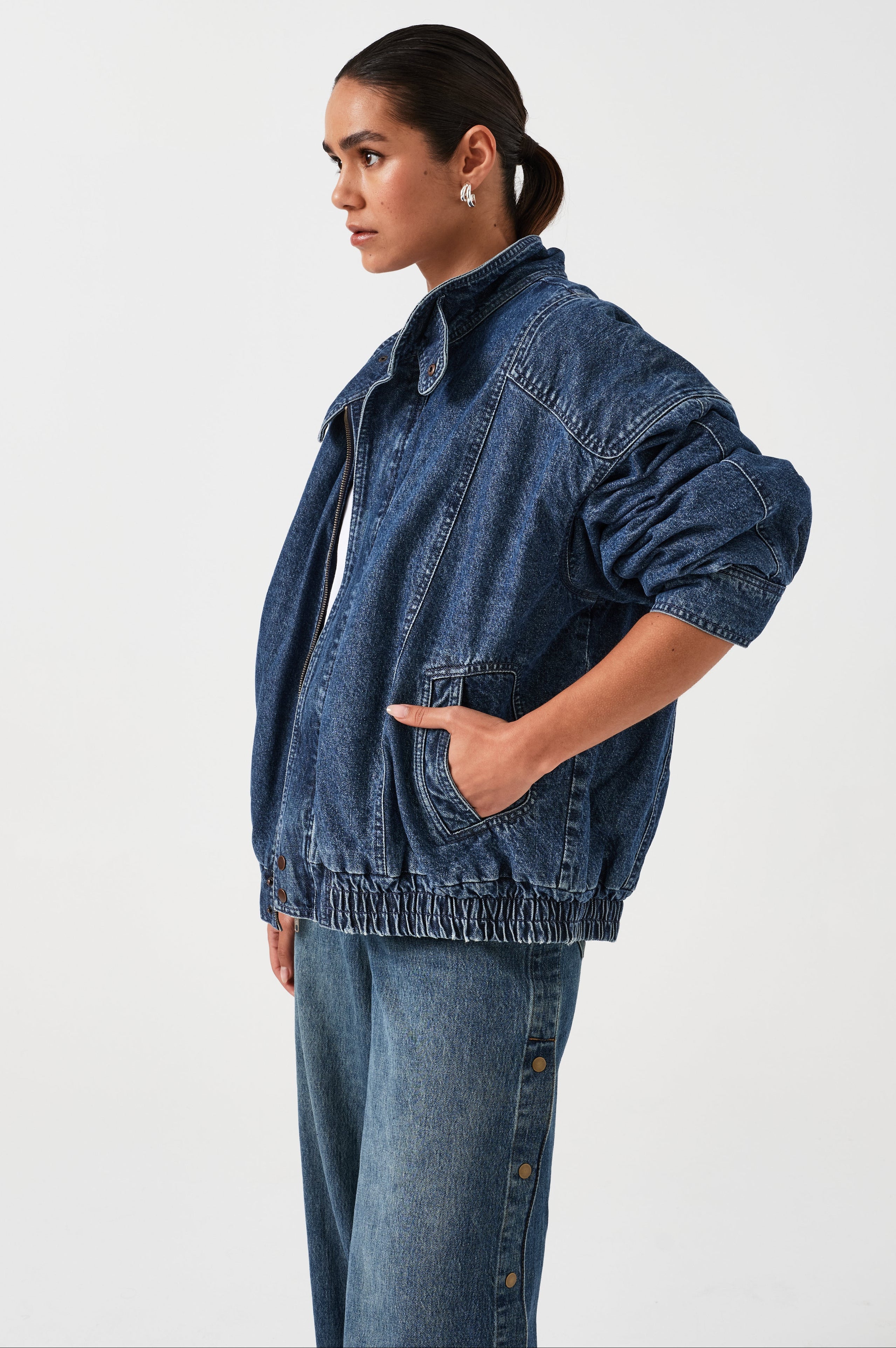 A woman wearing an oversized padded and seamed blue denim retro style bomber jacket with a round neckline, zip, and button closures at the top, bottom, and collar with a white top and blue jeans against a white backdrop