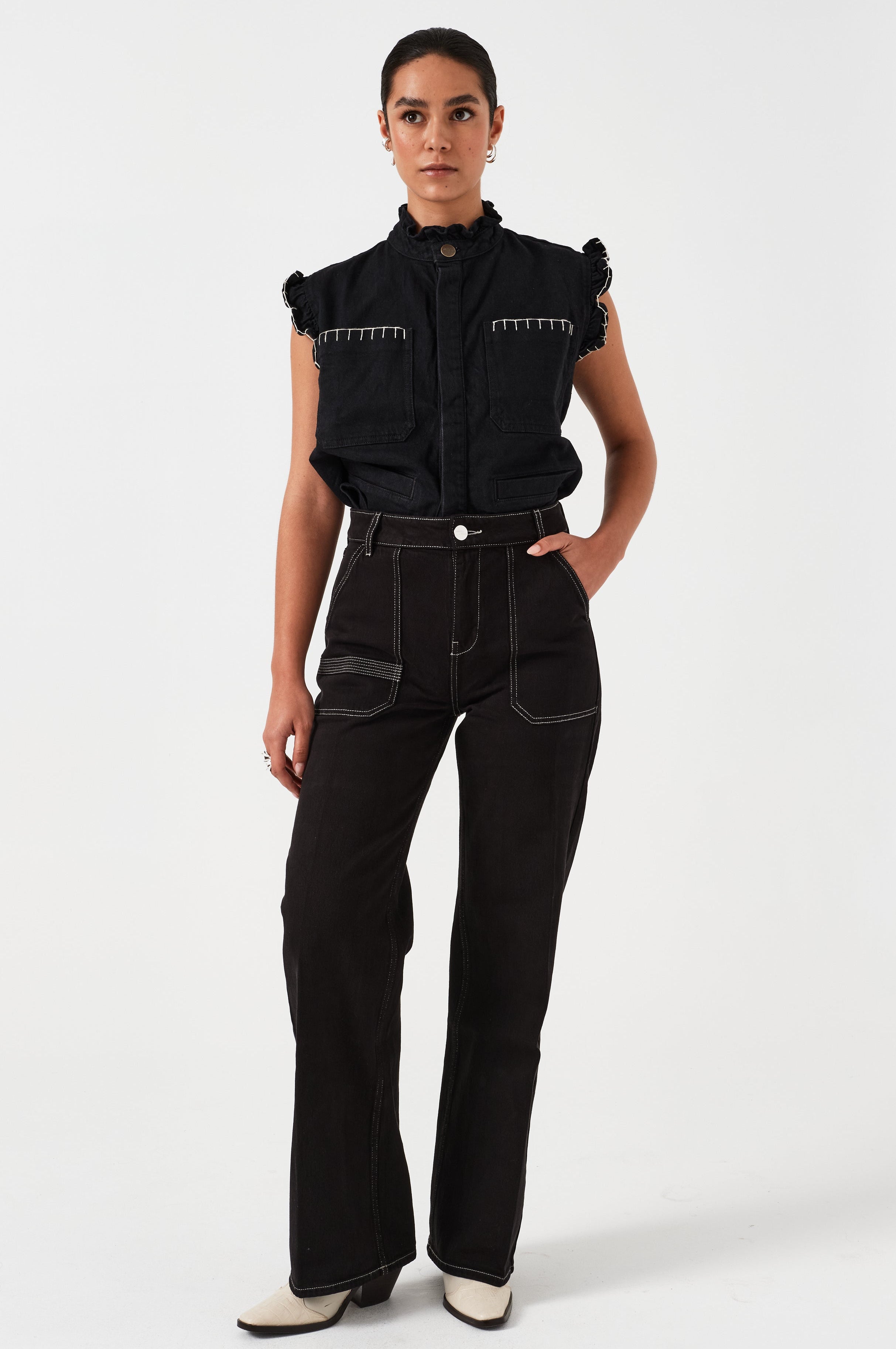 woman wearing a black vest/ waistcoat with a frill collar and our signature white stitching on two large patch pockets, paired with black jeans against a white backdrop