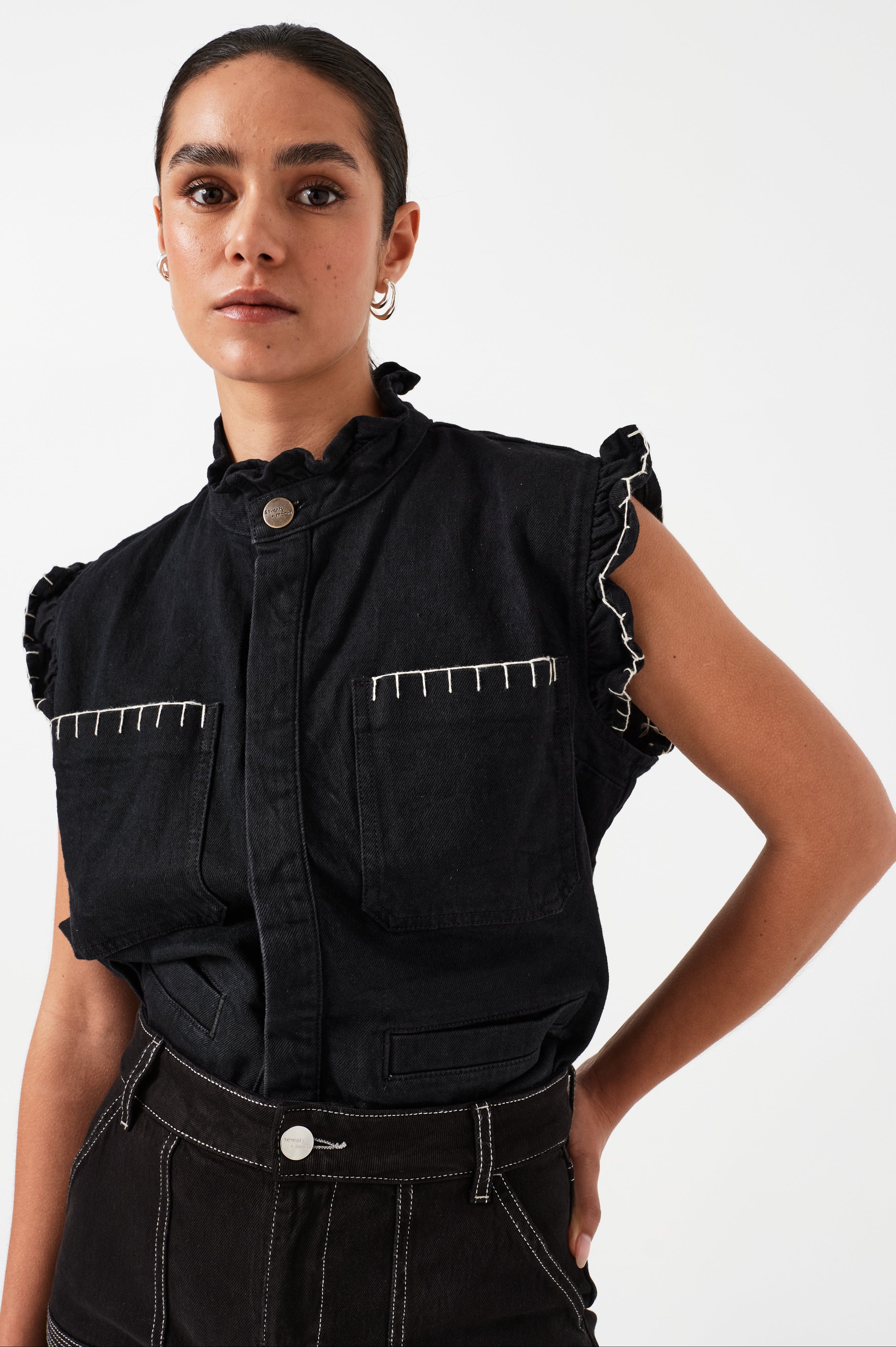 woman wearing a black vest/ waistcoat with a frill collar and our signature white stitching on two large patch pockets, paired with black jeans against a white backdrop