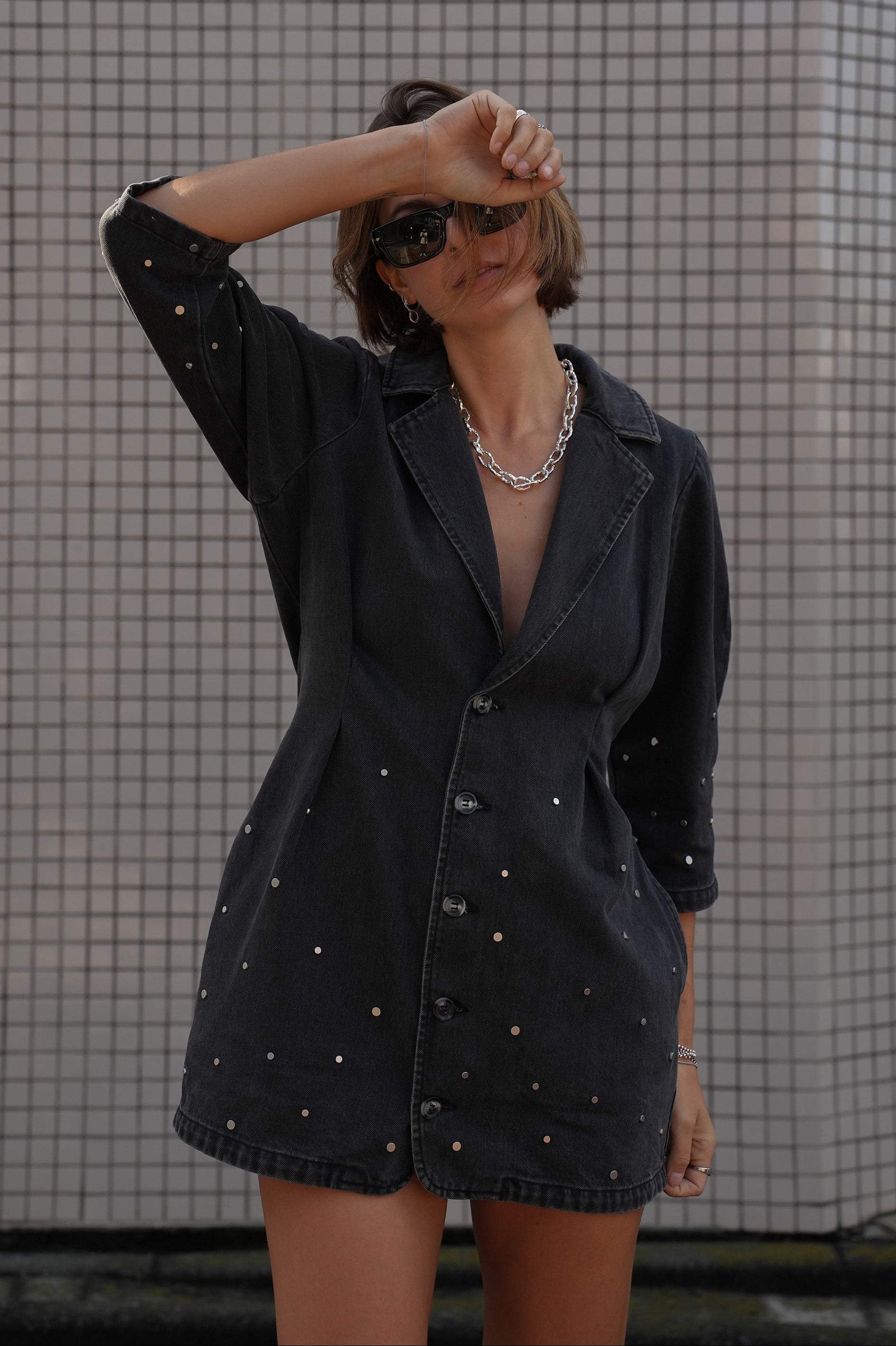 Woman wearing a black denim dress with silver studs (embellishments) and a tailored cinched waist against a tiled wall.