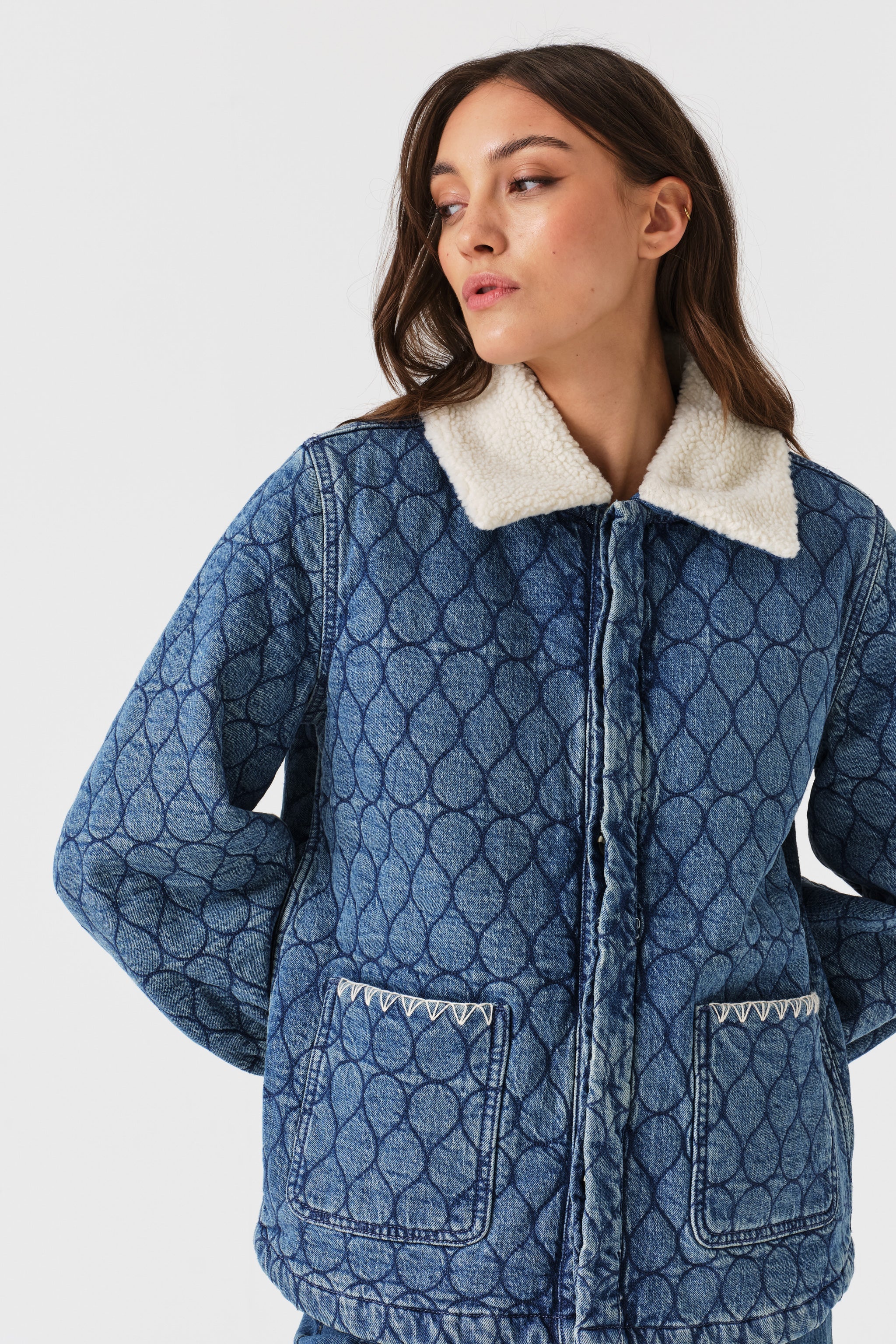 Woman wearing a blue quilted denim jacket with a removable white sherpa collar, large front pockets, and white embroidery details on a white background