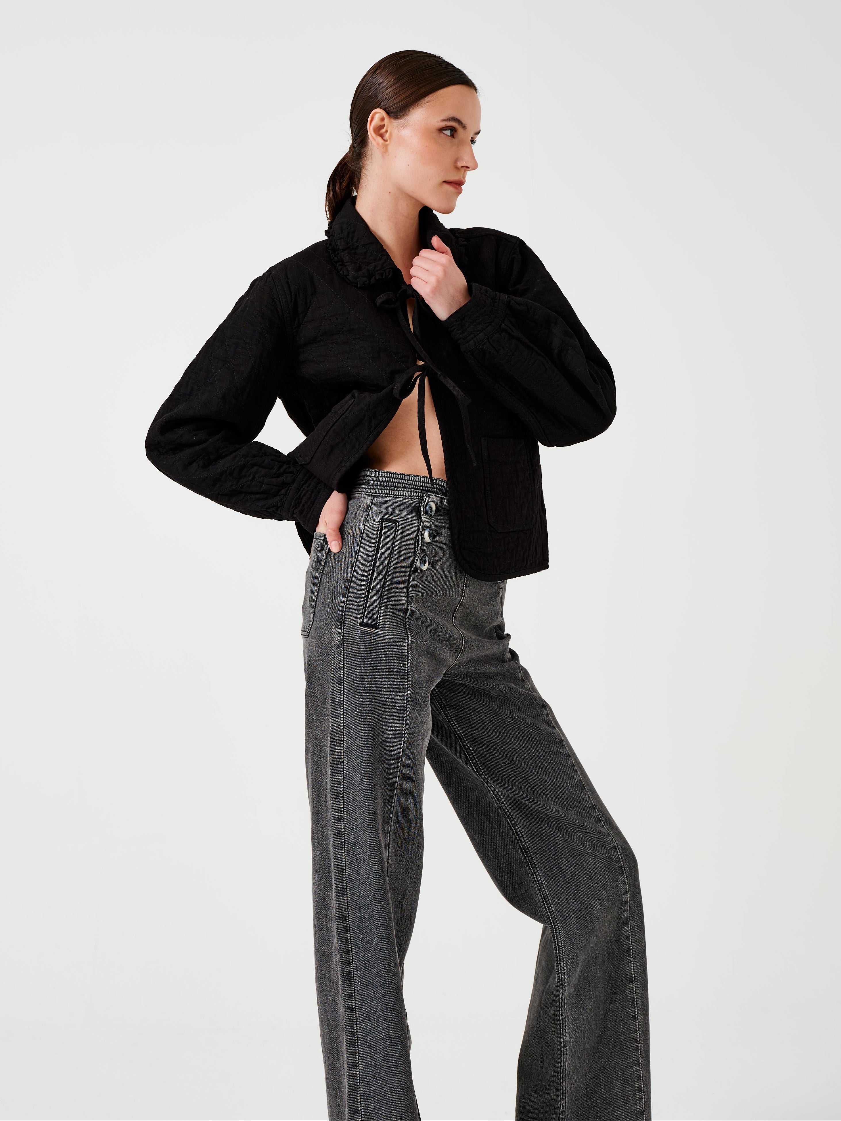 Woman wearing a black quilted denim jacket with a ruffled collar, front tie closures, and two large pockets, and black jeans on a white background