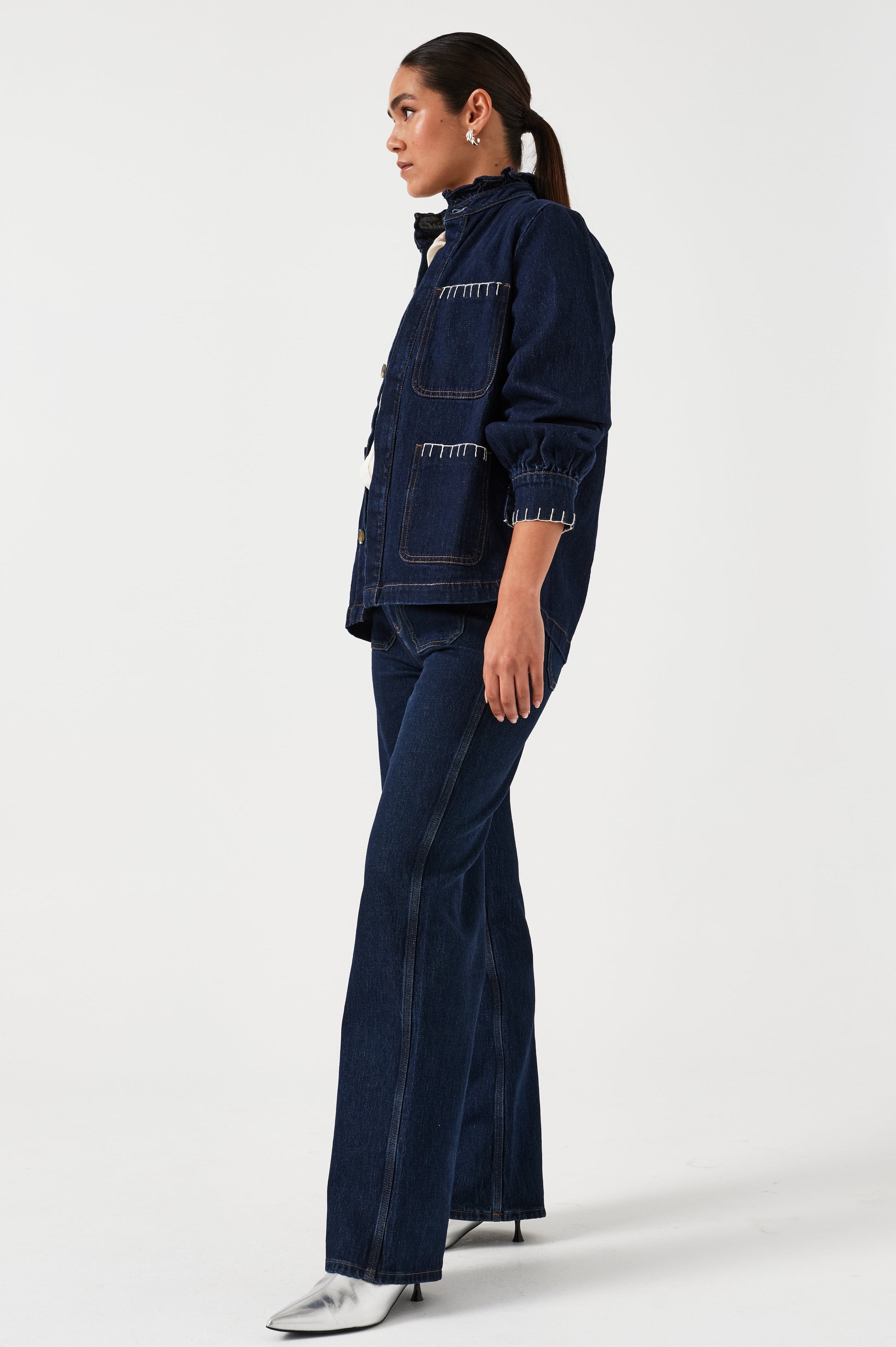 A woman wearing a dark blue vintage style jacket with a frill collar and our signature white blanket stitching on four large patch pockets, paired with a dark blue jeans and a white top against a white background