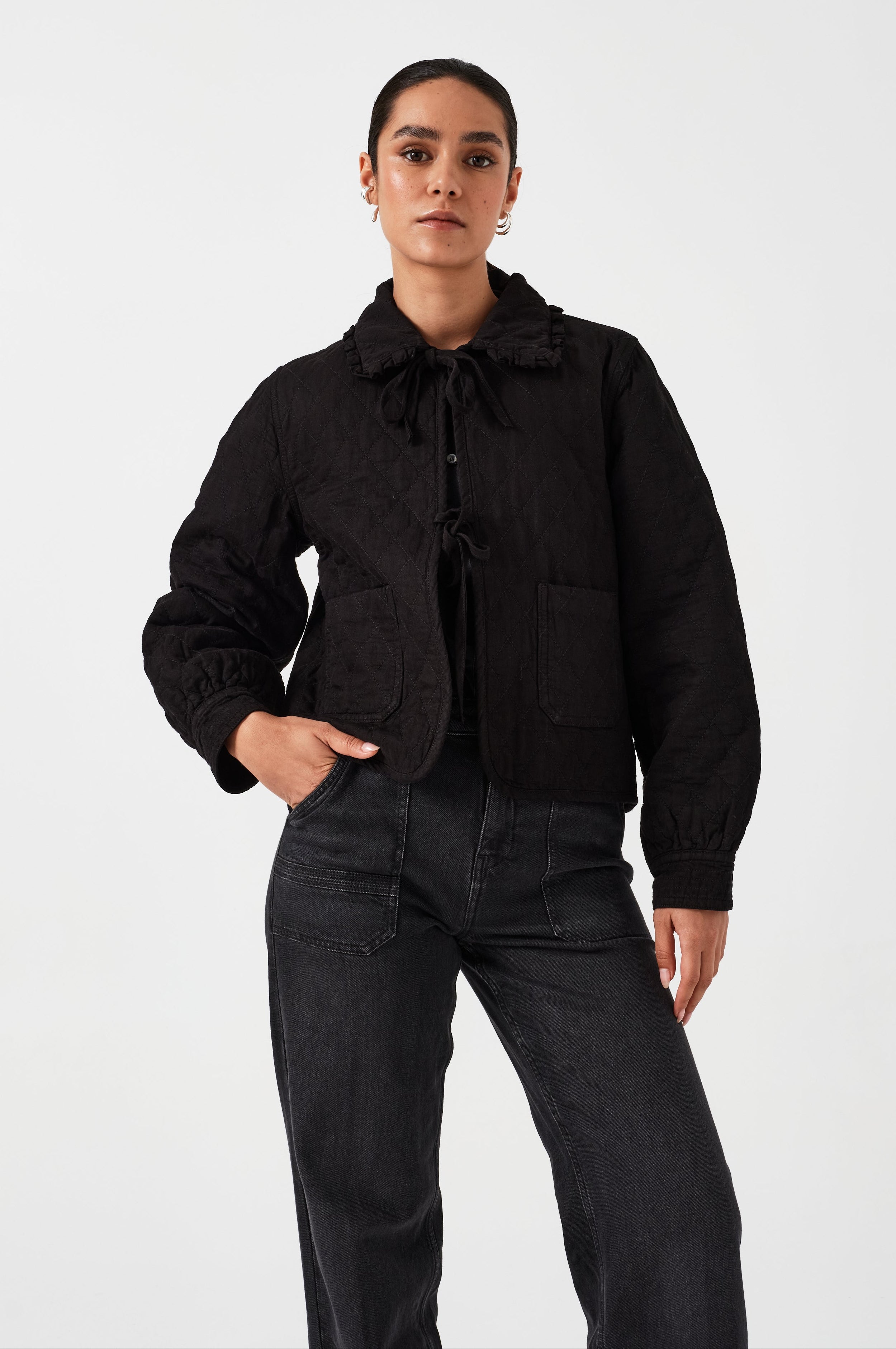 Woman wearing a black quilted denim jacket with a ruffled collar, front tie closures, and two large pockets on a white background