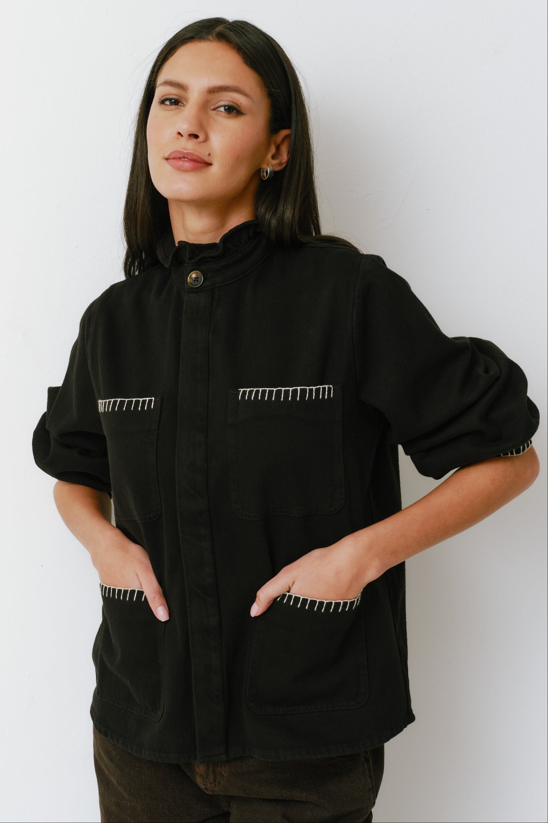 A woman wearing a black denim jacket with a frill collar and our signature white blanket stitching on four large patch pockets, paired with brown jeans against a white backdrop