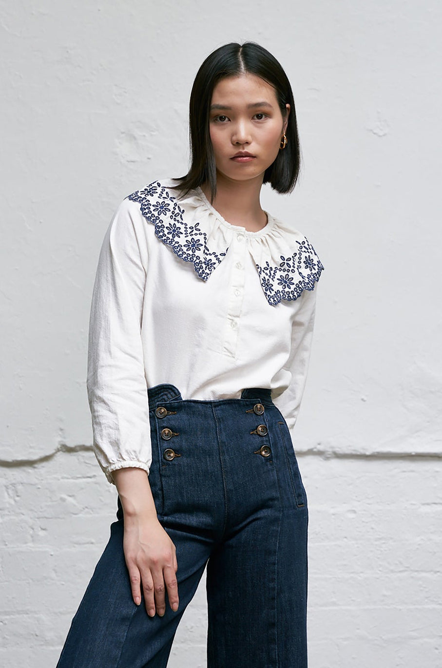 Woman wearing a white blouse with blue floral embroidery details on an eyelet style collar with blue jeans against a white background