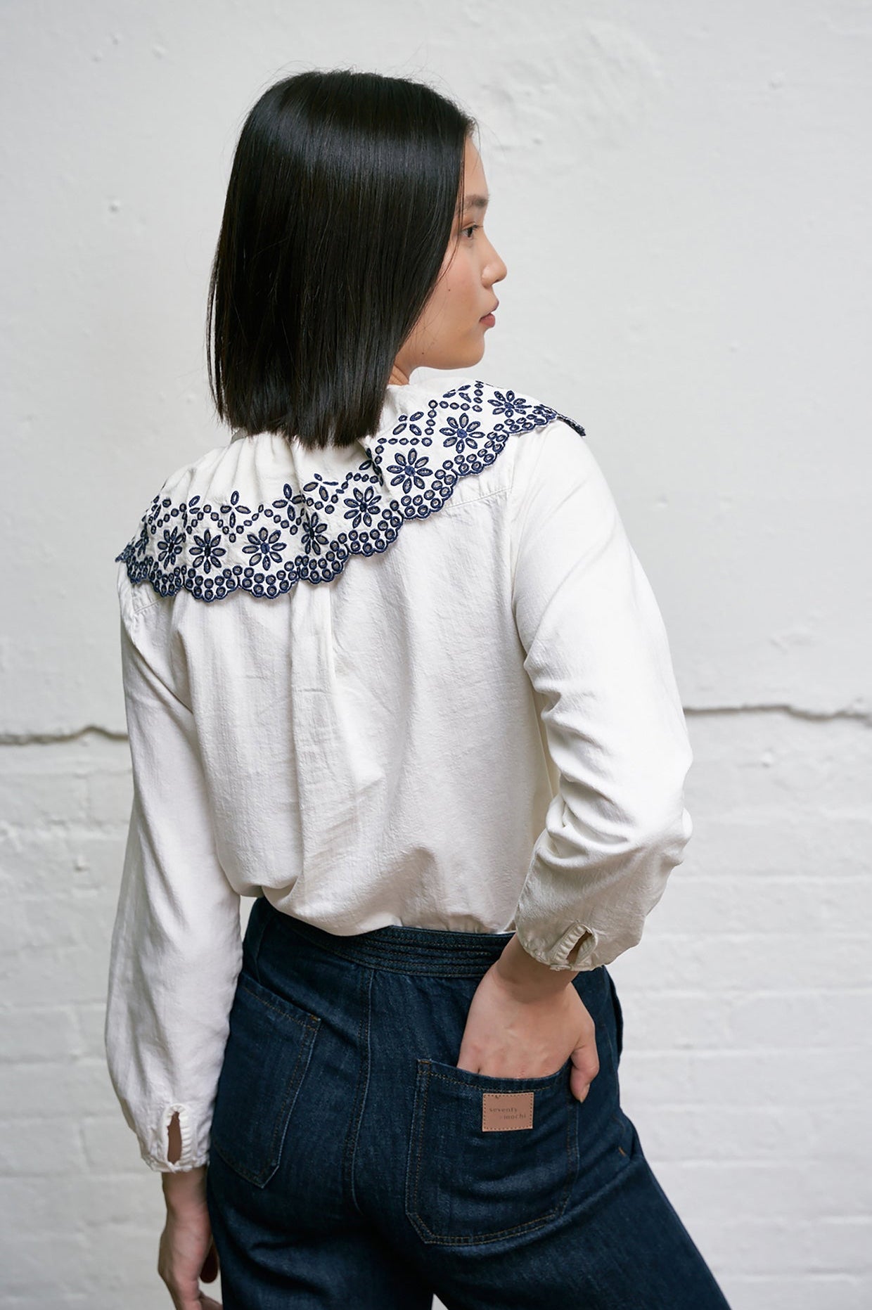 Woman wearing a white blouse with blue floral embroidery details on an eyelet style collar with blue jeans against a white background