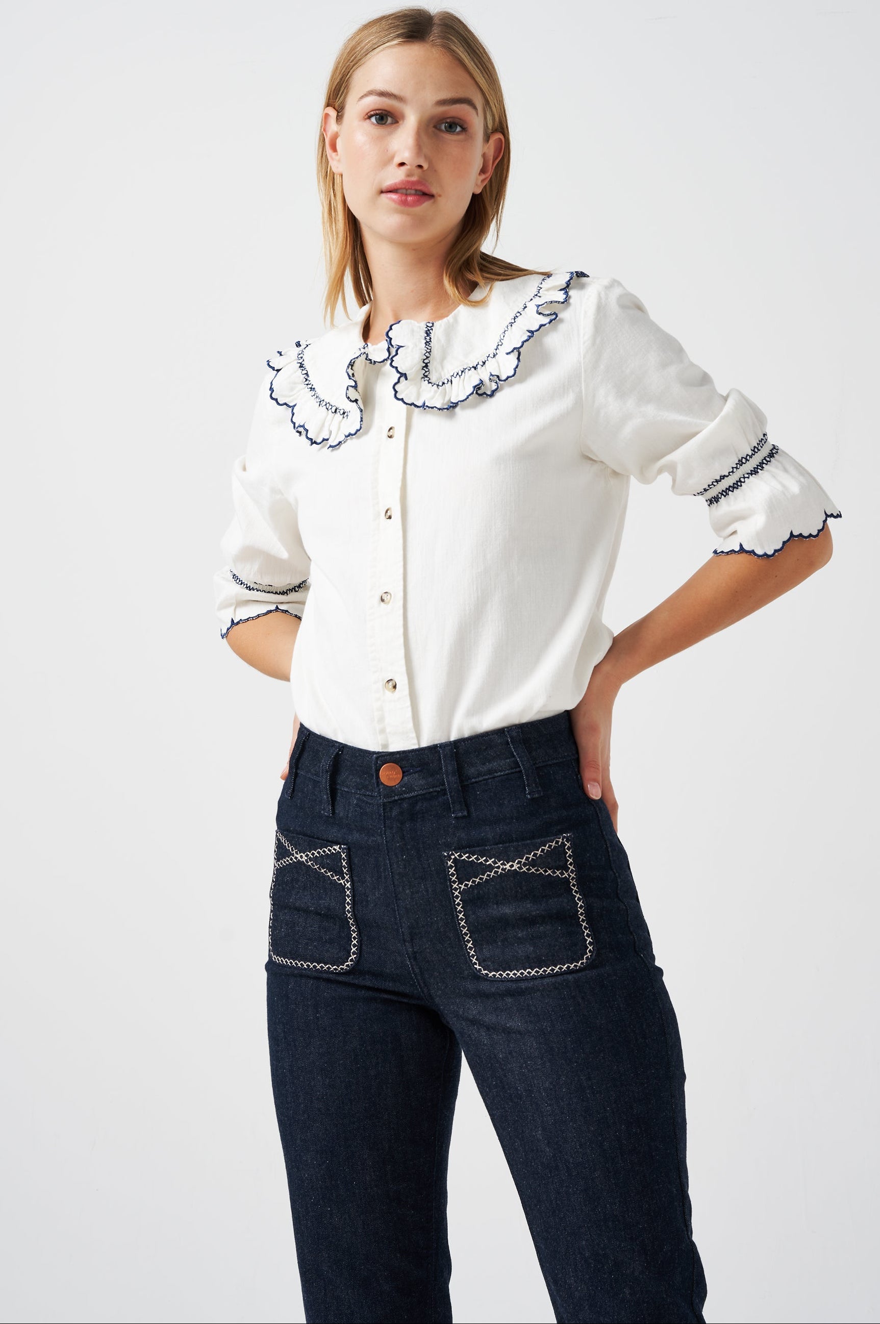 A woman wearing a white organic cotton blouse with dark blue embroiderery on frilled sleeves and a frill peek-a-boo collar with dark blue jeans on a white background