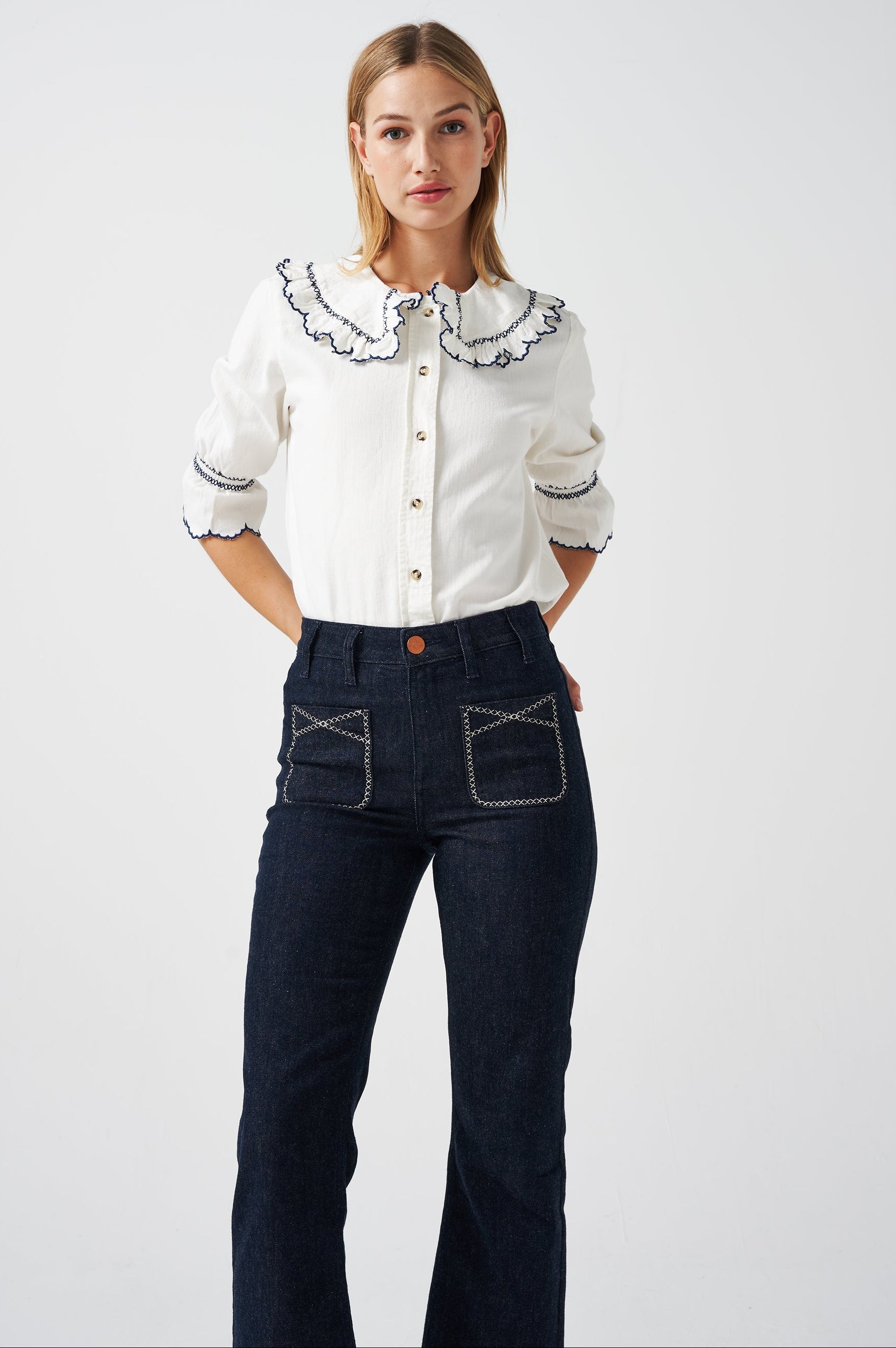 A woman wearing a white organic cotton blouse with dark blue embroiderery on frilled sleeves and a frill peek-a-boo collar with dark blue jeans on a white background