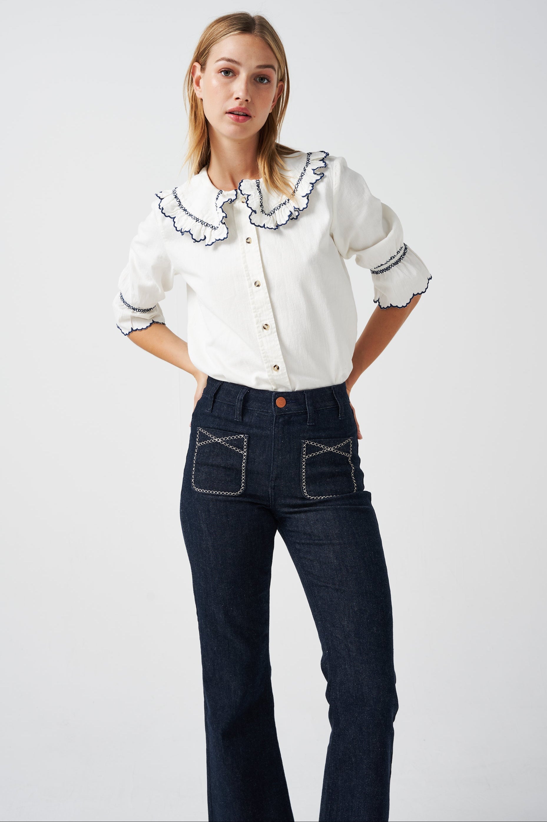 A woman wearing a white organic cotton blouse with dark blue embroiderery on frilled sleeves and a frill peek-a-boo collar with dark blue jeans on a white background