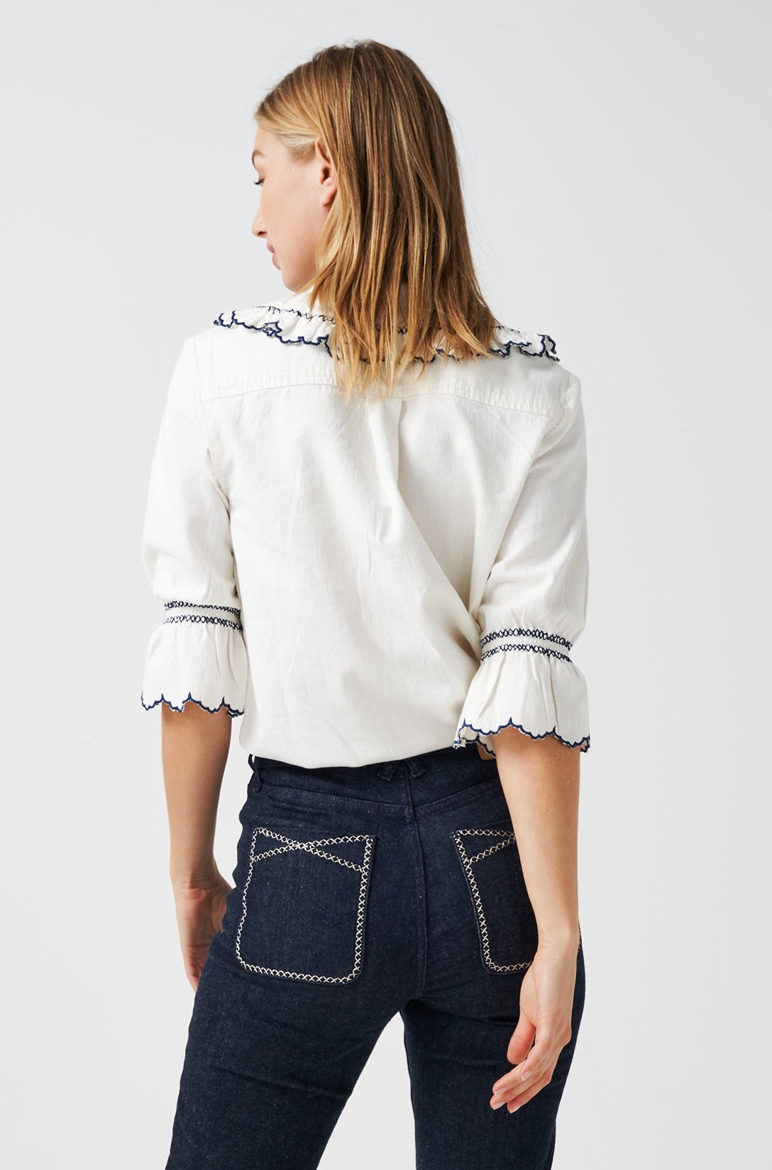 A woman wearing a white organic cotton blouse with dark blue embroiderery on frilled sleeves and a frill peek-a-boo collar with dark blue jeans on a white background