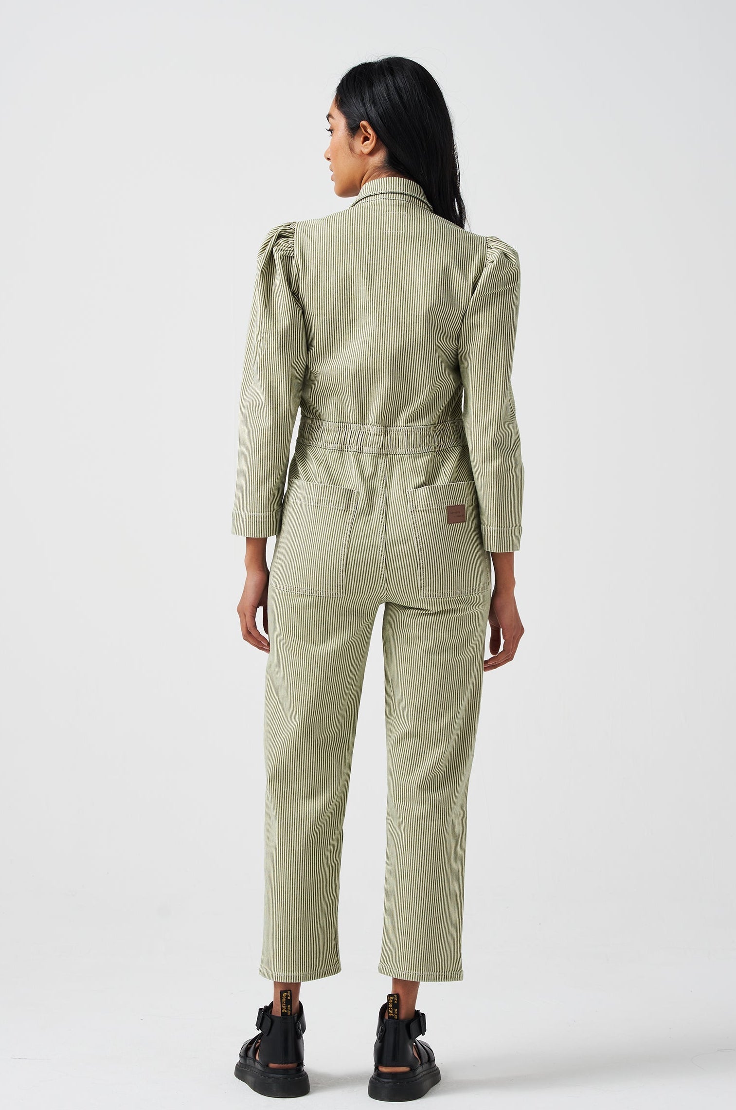 A woman wearing a khaki green and white striped jumpsuit with soft puff shoulders, large pockets, and a front zip detail on a white background