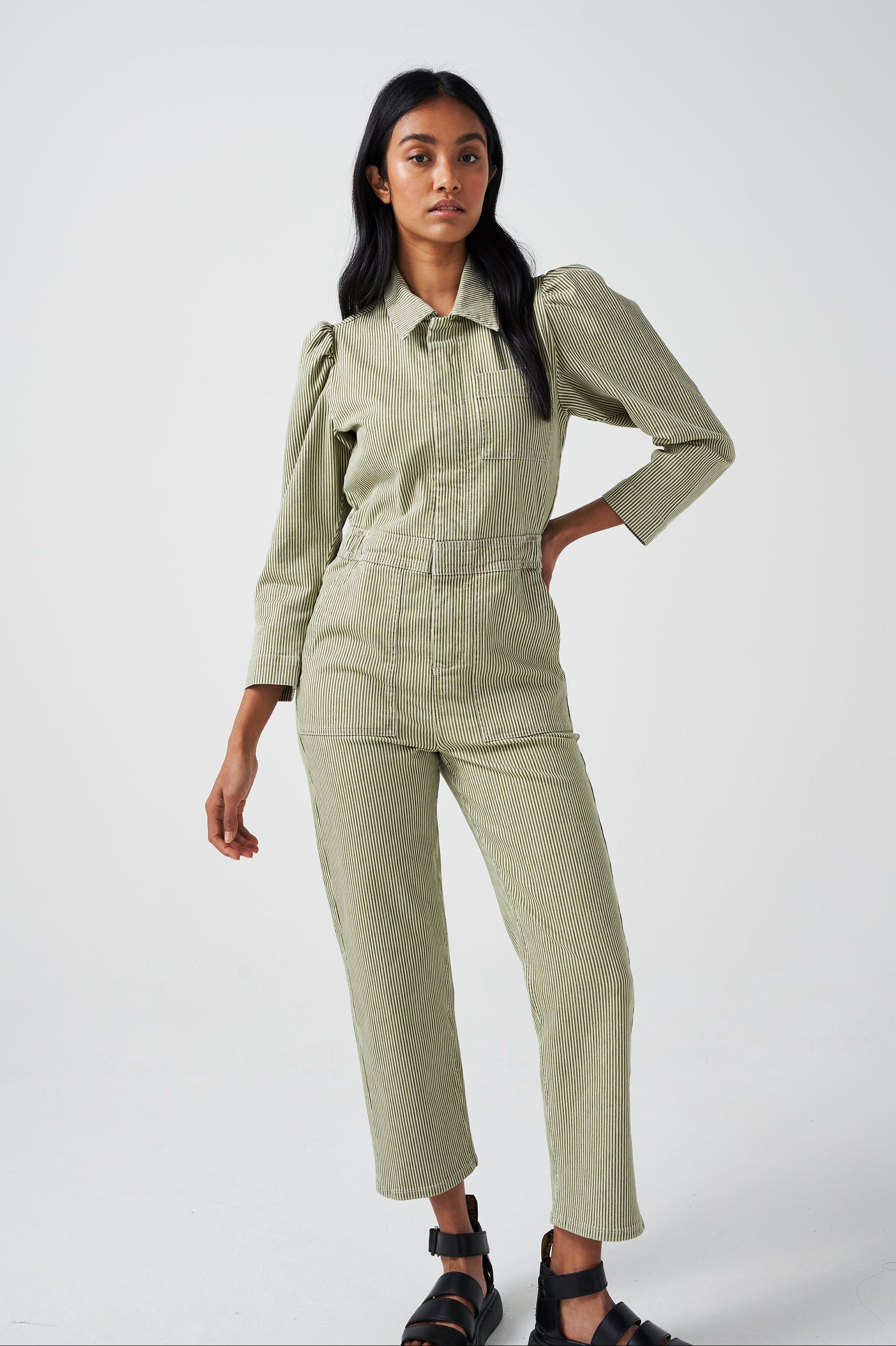 A woman wearing a khaki green and white striped jumpsuit with soft puff shoulders, large pockets, and a front zip detail on a white background