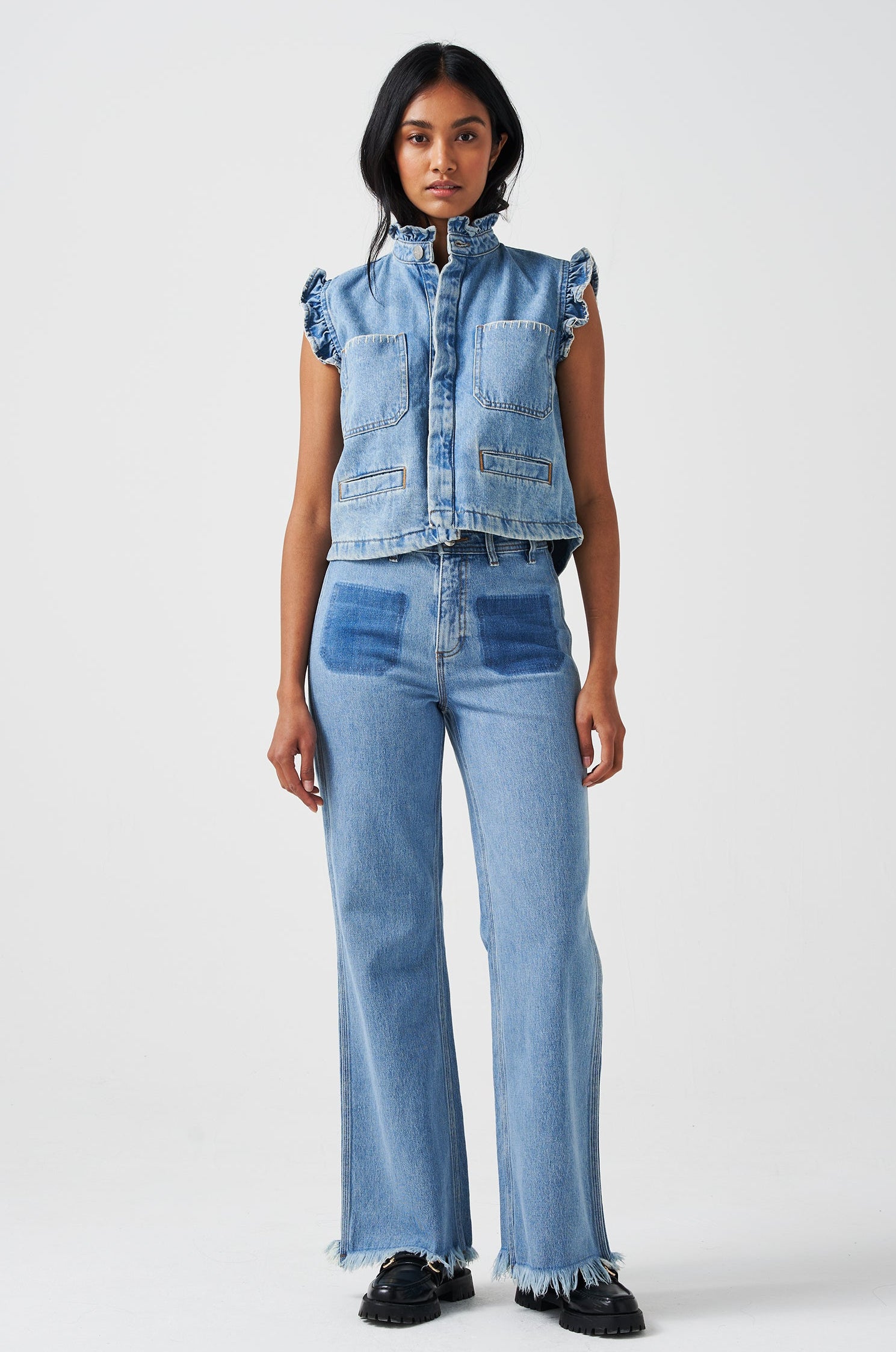 woman wearing a blue denim waistcoat/ vest with frill collar and sleeves, and two large patch pockets with white stitching, paired with blue jeans on a white background