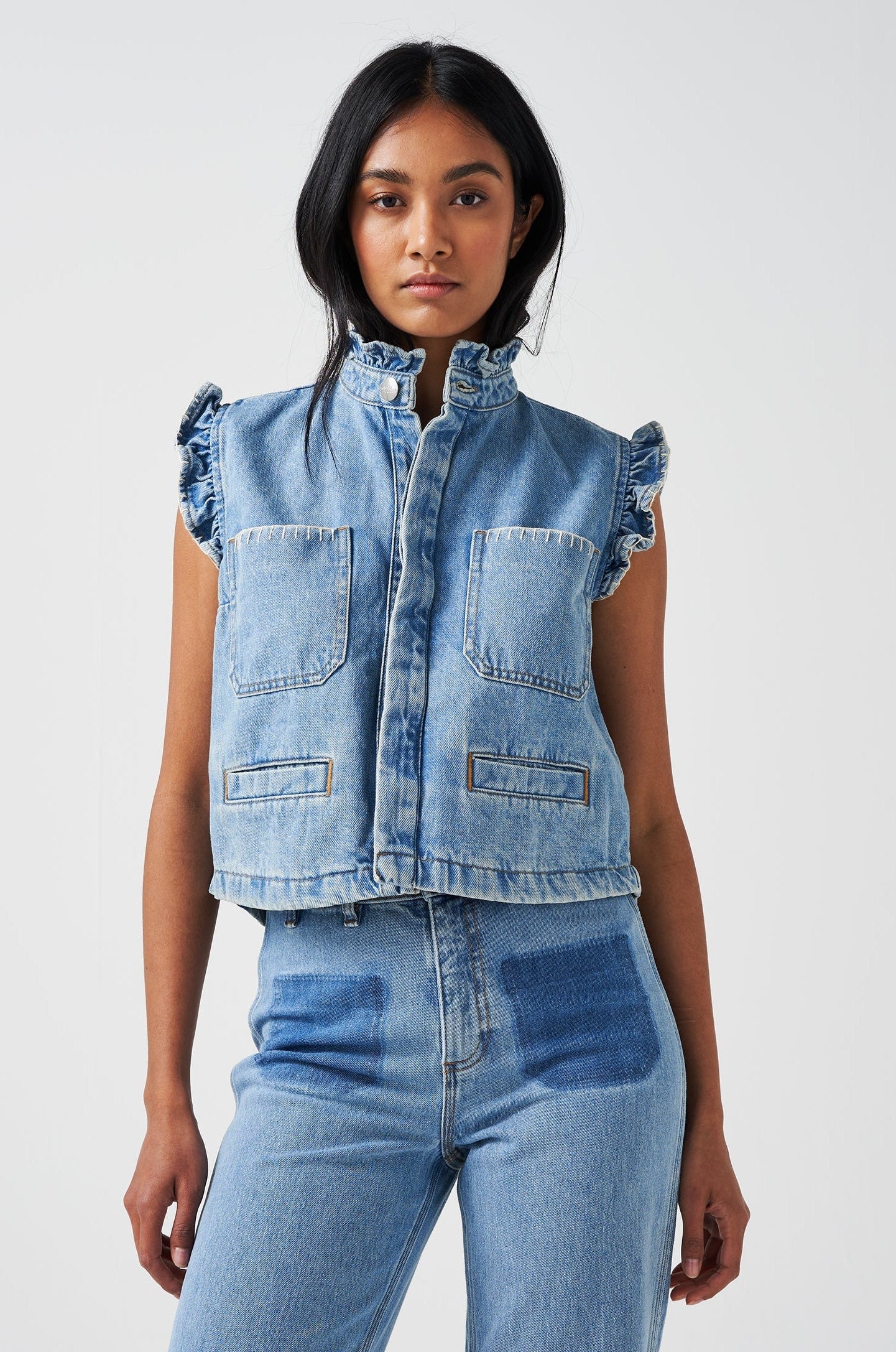 woman wearing a blue denim waistcoat/ vest with frill collar and sleeves, and two large patch pockets with white stitching, paired with blue jeans on a white background