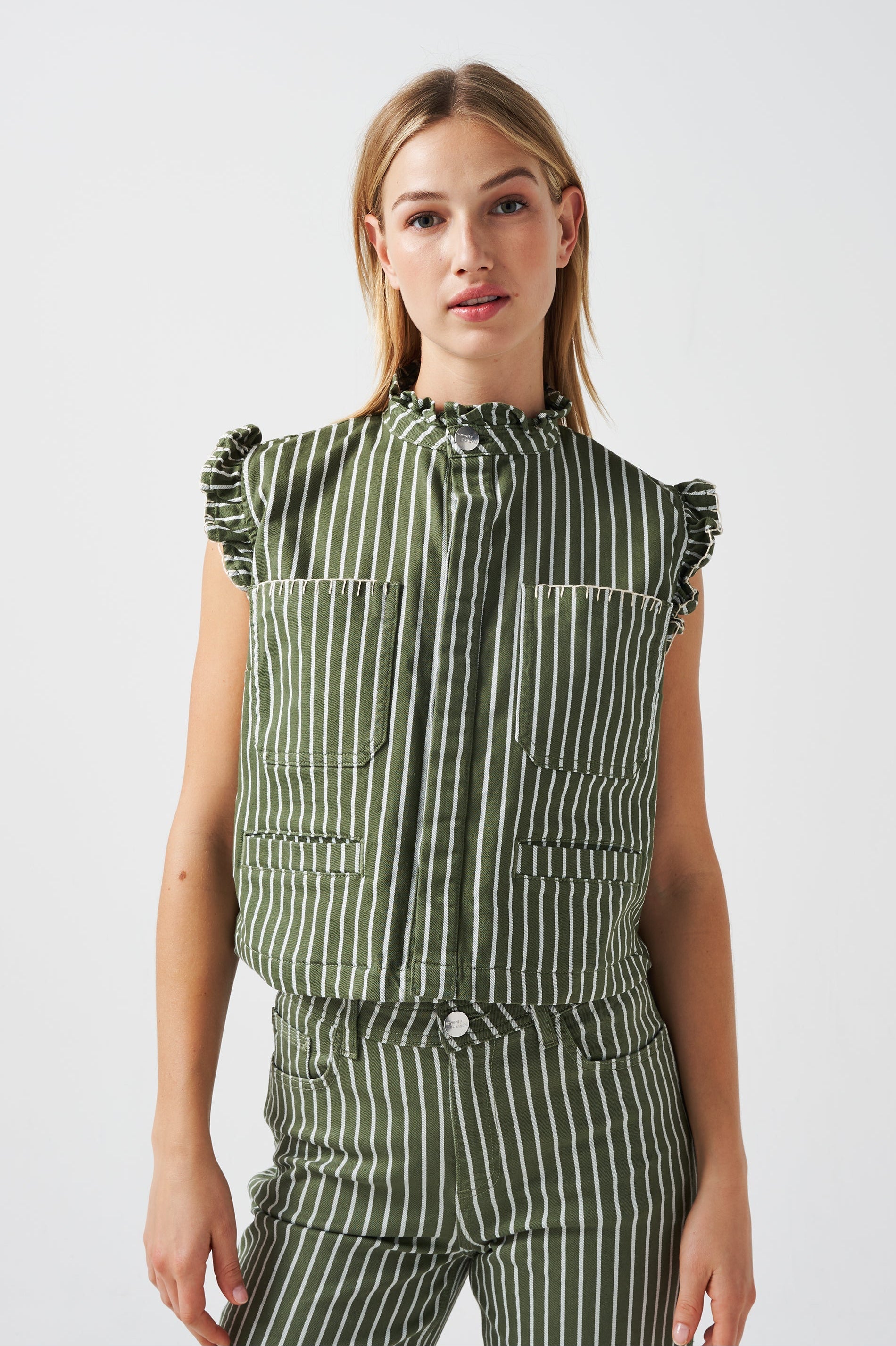 A woman wearing a khaki and white striped vest/ waistcoat with a frill collar, white blanket stitching, and two large patch pockets, part of a matching set, on a white background.