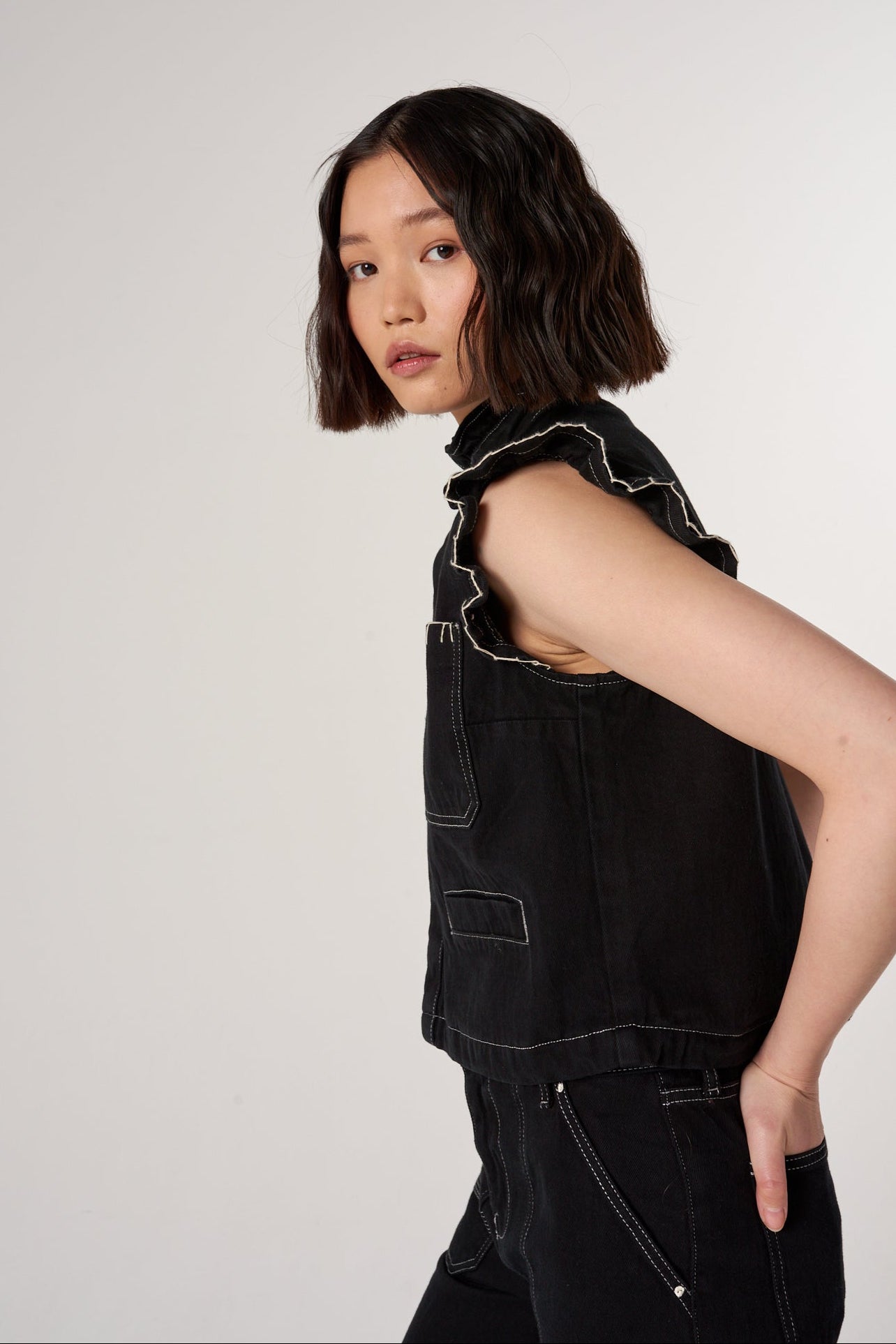A woman wearing a black denim vest / waistcoat with a frill collar, white blanket stitch details, and two large patch pockets, paired with black denim trousers against a white background